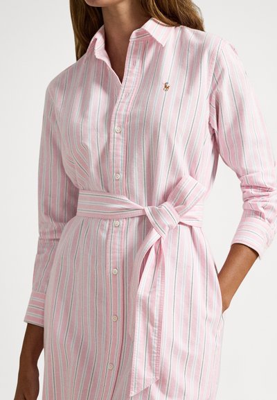 Sieviete, tērpusies rozā un baltā svītraina pogājama kleitā ar sasienamu jostas siksnu viduklī un nelielu izšuvumu ar logotipu uz krūtīm.