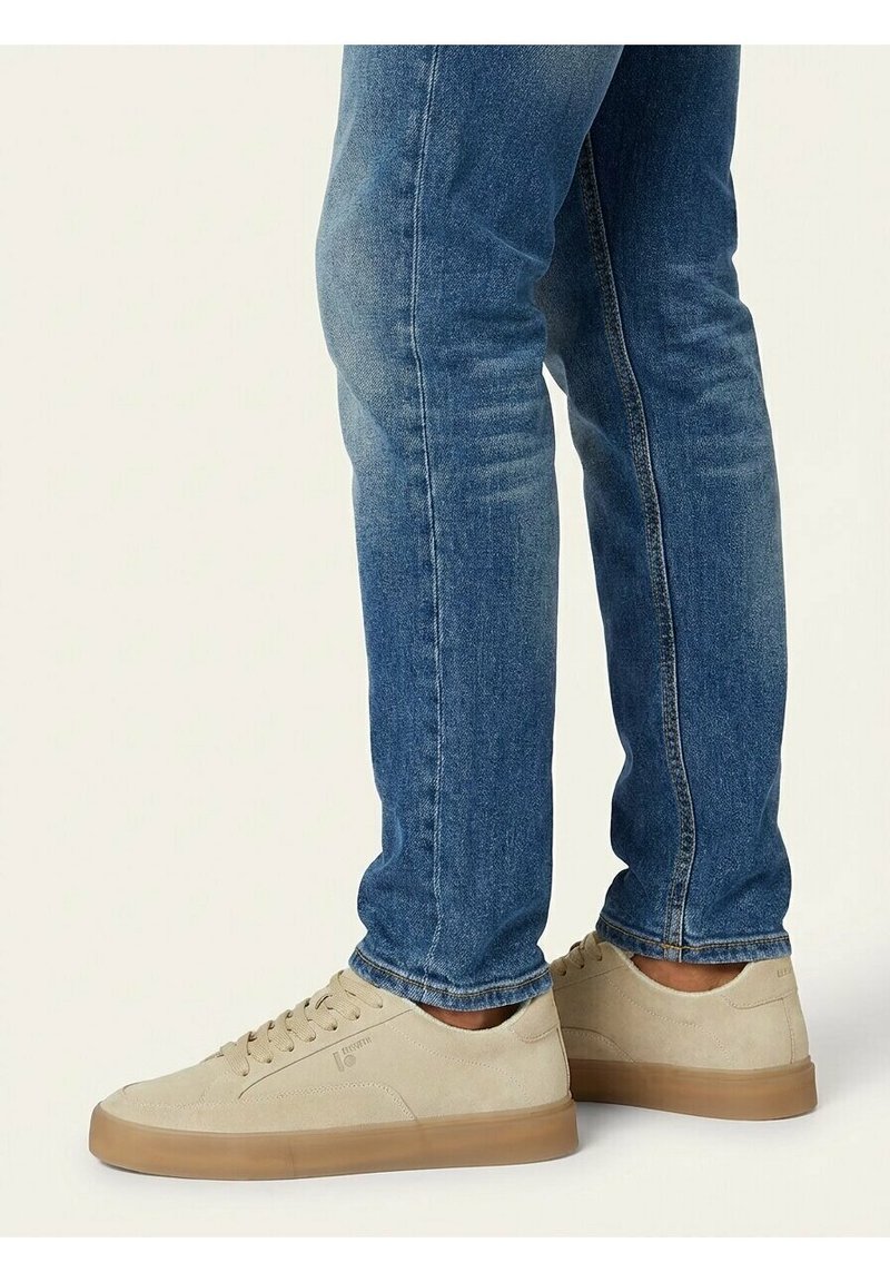 Persoon draagt een blauwe spijkerbroek en beige suède sneakers met gumzolen, staand op een effen lichte achtergrond.