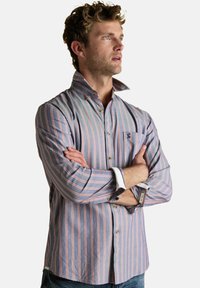 Vyras su garbanotais plaukais, dėvintis mėlynai ir rožine juostuotą marškinėlį su apykakle, sukryžiuotomis rankomis, žiūri į dešinę ant baltos fono.