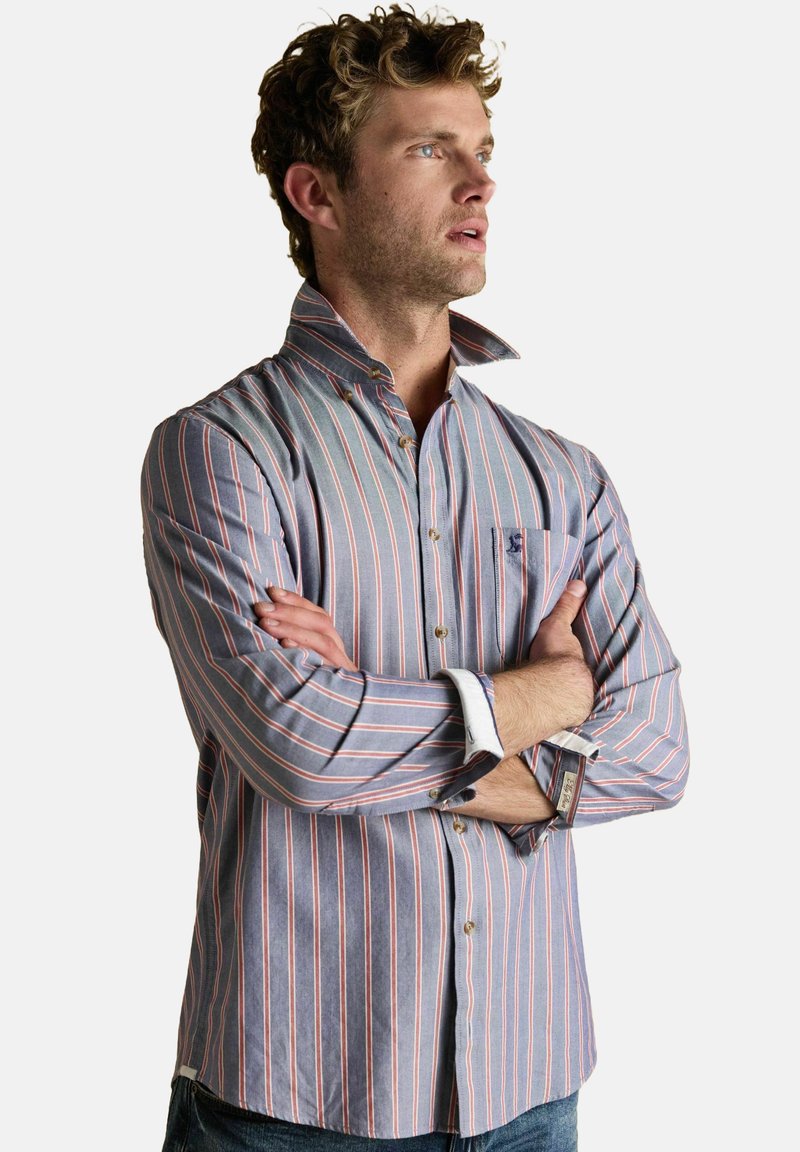 Vyras su garbanotais plaukais, dėvintis mėlynai ir rožine juostuotą marškinėlį su apykakle, sukryžiuotomis rankomis, žiūri į dešinę ant baltos fono.