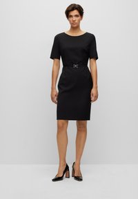 Black fitted dress with short sleeves, round neckline, and waist belt featuring a metallic buckle. Model wearing black high-heeled shoes.