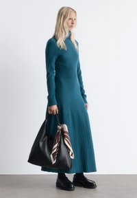 Frau in einem türkisfarbenen, plissierten Langarmkleid, die eine große schwarze Lederhandtasche hält, dekoriert mit einem gemusterten Seidenschal, und schwarze Plateauschuhe trägt.