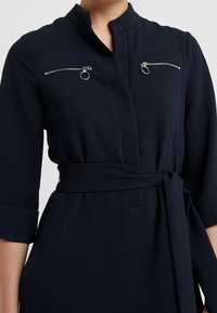 Granatowa sukienka z wysokim kołnierzem, długimi rękawami z mankietami, dwiema kieszeniami zapinanymi na zamek na klatce piersiowej oraz paskiem w talii.