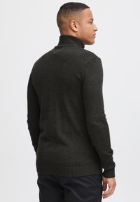 Suéter de punto verde oscuro con cuello alto y puños acanalados. Presenta una textura suave y un diseño ajustado. Vista trasera, usado por una persona.