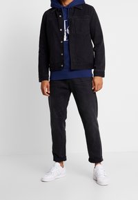 Giacca di jeans nera sopra un hoodie blu navy, abbinata a jeans scuri e sneakers bianche. Il look presenta bottoni e maniche arrotolate.