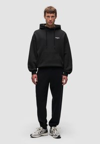 Sudadera negra de gran tamaño con bolsillo frontal, capucha con cordón y logotipo blanco. Combinada con joggers negros ajustados y zapatillas reflectantes.