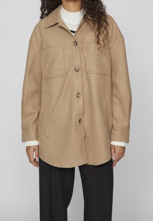 Personne portant un manteau en laine beige boutonné avec deux poches poitrine, par-dessus un pull blanc et un pantalon noir, sur un fond uni.
