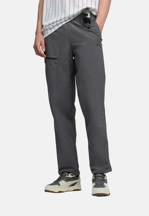 Persona con pantalones cargo gris oscuro con bolsillos y zapatillas negras y blancas, de pie con una mano en el bolsillo.