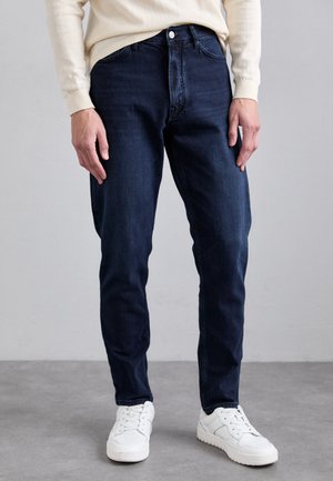 Mörkblå denimjeans med rak passform, femfickorsstil och knappslå. Bärs ihop med vita sneakers och en ljusfärgad tröja.