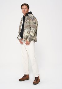 Kurtka w wzór camouflage z czarną bluzą z kapturem pod spodem, białe spodnie i brązowe buty sznurowane. Charakteryzuje się różnorodnymi odcieniami zieleni i brązu.