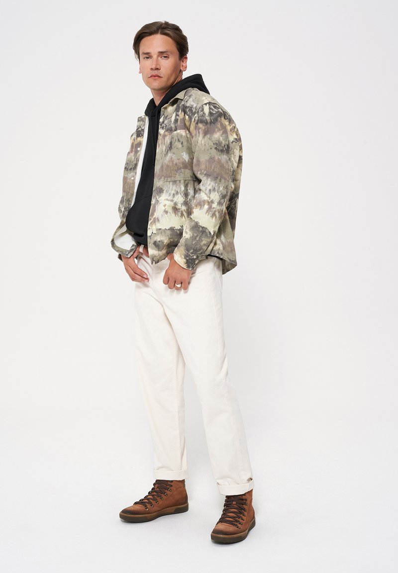 Kurtka w wzór camouflage z czarną bluzą z kapturem pod spodem, białe spodnie i brązowe buty sznurowane. Charakteryzuje się różnorodnymi odcieniami zieleni i brązu.