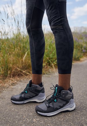 Vysoké atletické boty s šedým a černým barevným schématem, vyznačující se tyrkysovými akcenty a texturovanými překryvy. Robustní design s tkaničkami.