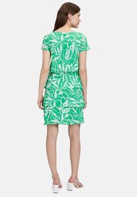 Grünes Kleid mit blättrigen weißen Mustern, kurzen Ärmeln und gestuftem Rüschensaum. Hergestellt aus leichtem Stoff, präsentiert sich in einem lässigen Schnitt.