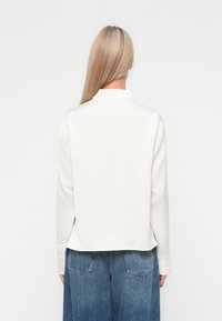 Hvid langærmet top med høj krave, der har en afslappet pasform og en rygplissédetalje, parret med løse blå denimjeans.