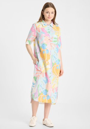 Młoda kobieta ubrana w pastelową, wielokolorową kwiatową sukienkę midi zapinaną na guziki, z krótkimi rękawami i białe trampki, stojąca z jedną ręką w kieszeni.