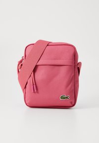 Rosa stoff kryssbodiesekk med en teksturert overflate, justerbar stropp og en liten grønn krokodillelogo på forsiden. Med glidelåslomme.
