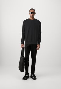 Felpa nera con logo in rilievo, pantaloni neri e borsa a tracolla nera trapuntata. Le calzature presentano un design testurizzato con dettagli bianchi.