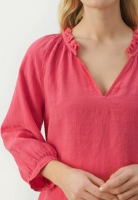 Roosa linane pluus V-kaeluse, volangiga kaeluse ja pikkade varrukatega, millel on kokku kokkuvõetud mansetid. Kangas näeb välja kerge ja pehme tekstuuriga.