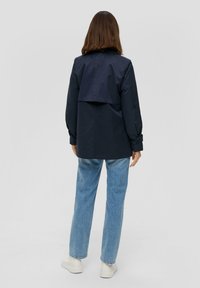 Marineblaue Jacke mit Rückenklappe, langen Ärmeln und knöpfbaren Manschetten, getragen über hellblauen Jeans und weißen Sneakers. Glatte Stoffoberfläche.