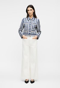 Nő, kék-fehér függőleges csíkos dzsekit arany gombokkal visel, fehér bőszárú nadrágot és fekete hegyes cipőt, zsebében tartott kézzel áll.