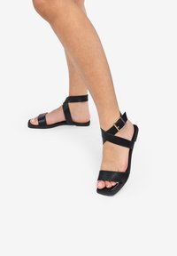 Sandalias de cuero negro con suela plana, con tiras entrelazadas y un acento de hebilla lateral. Diseño de punta abierta, minimalista y elegante.