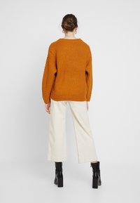 Übergroßer, gestrickter Pullover in rostfarben mit weitem Halsausschnitt, strukturiertem Muster und tiefen Schultern, kombiniert mit cremefarbener, weit geschnittenen Hose und schwarzen Stiefeln.