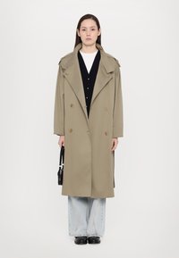 Olive green trench coat with oversized lapels, double-breasted front, and shoulder details. Paired with black cardigan and light blue flared jeans.