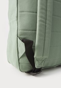 Mochila de lona verde con textura acolchada, correas de hombro acolchadas y un bolsillo lateral con una correa ajustable negra.