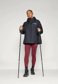 Vrouw in een zwarte hooded jacket en rode legging, met wandelstokken in de hand, staand tegen een effen achtergrond.