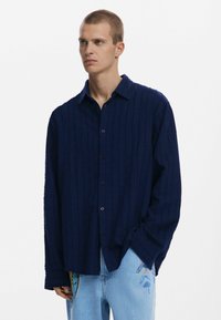 Marineblå skjorte med knapper og teksturerede striber, lange ærmer og en afslappet pasform. Kombineret med lyseblå jeans med farverige accenter.