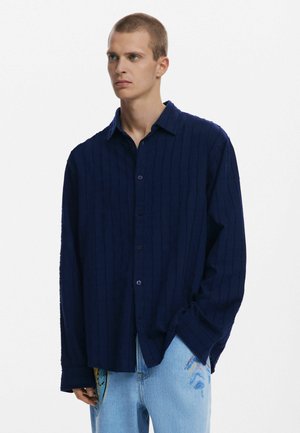 Navy blue button-up shirt with textured stripes, long sleeves, and a relaxed fit. Paired with light blue jeans featuring colorful accents.