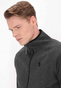 Giacca grigia con zip e colletto alto, realizzata in tessuto liscio, con un logo nero sul lato sinistro del petto e un dettaglio della cerniera metallicoa.