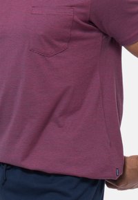 Bordeaux gestreepte t-shirt met een borstzak, gedragen door een persoon naast donkere broek, waarbij de zijkant en stiksels van het shirt worden getoond.