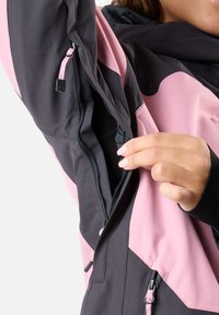 Schwarze und rosa Jacke mit einer Reißverschlusstasche, glatter Textur und kontrastierenden Farbblöcken. Verstellbarer Zugring am Reißverschluss.