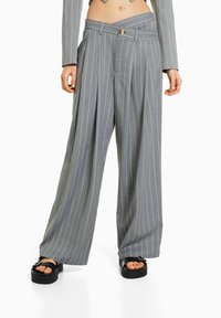 Grijze high-waisted pantalon met finestripes en brede pijpen, met een overlappend tailleband, gedragen met zwarte platform sandalen, waardoor de blote buik zichtbaar is.