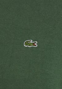 Grönstickad tröja med en liten broderad krokodillogo med röda detaljer på vänster sida. Mjuk textur och figursydd design.