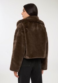 Giacca corta in finto pelo marrone con ampio colletto, texture morbida e maniche lunghe, abbinata a pantaloni in denim neri.