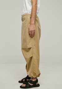 Beige bomuldsbukser med en afslappet pasform, der har elastiske manchetter og sidelommer, parret med sorte sandal-straps.