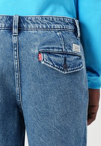 Light blue denim jeans featuring a back-pocket design, a red tab, and stitch detailing. The waistband has a branded tag.