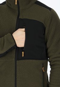 Giacca in pile verde oliva con pannello in tessuto nero, tasca a zip sul petto e pile testurizzato. Presenta un cursore della zip con accento beige.