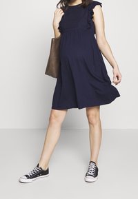 Marinstor blommig gravidklänning med volangärmar, texturerad bodice, flödande kjol och bärs med svarta sneakers. Brun toffeväska i handen.