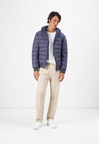 Young man wearing a navy blue puffer jacket, white t-shirt, beige trousers, and white sneakers, standing against a plain white background.