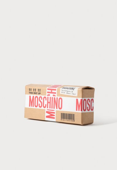 Boîte en carton rectangulaire scellée avec un ruban blanc imprimé « MOSCHINO » en rouge, affichant une étiquette d'adresse et un code-barres sur fond uni.