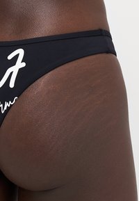 Juodas bikini dugnas, pagamintas iš tampraus audinio, su balta grafika šone. Lygi tekstūra su klasikiniu kirpimu.