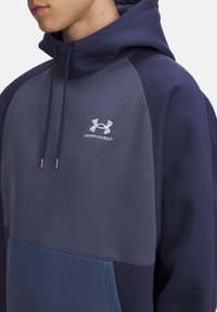 Mørkeblå hættebluse lavet af blødt stof, med et farveblokdesign med grå og lysere blå områder, samt et lille hvidt logo på brystet.