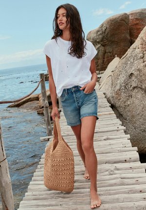 Femeie în top alb cu mâneci scurte și pantaloni scurți din denim, purtând o geantă împletită, mergând desculță pe o platformă de lemn lângă malul stâncos și marea calmă.