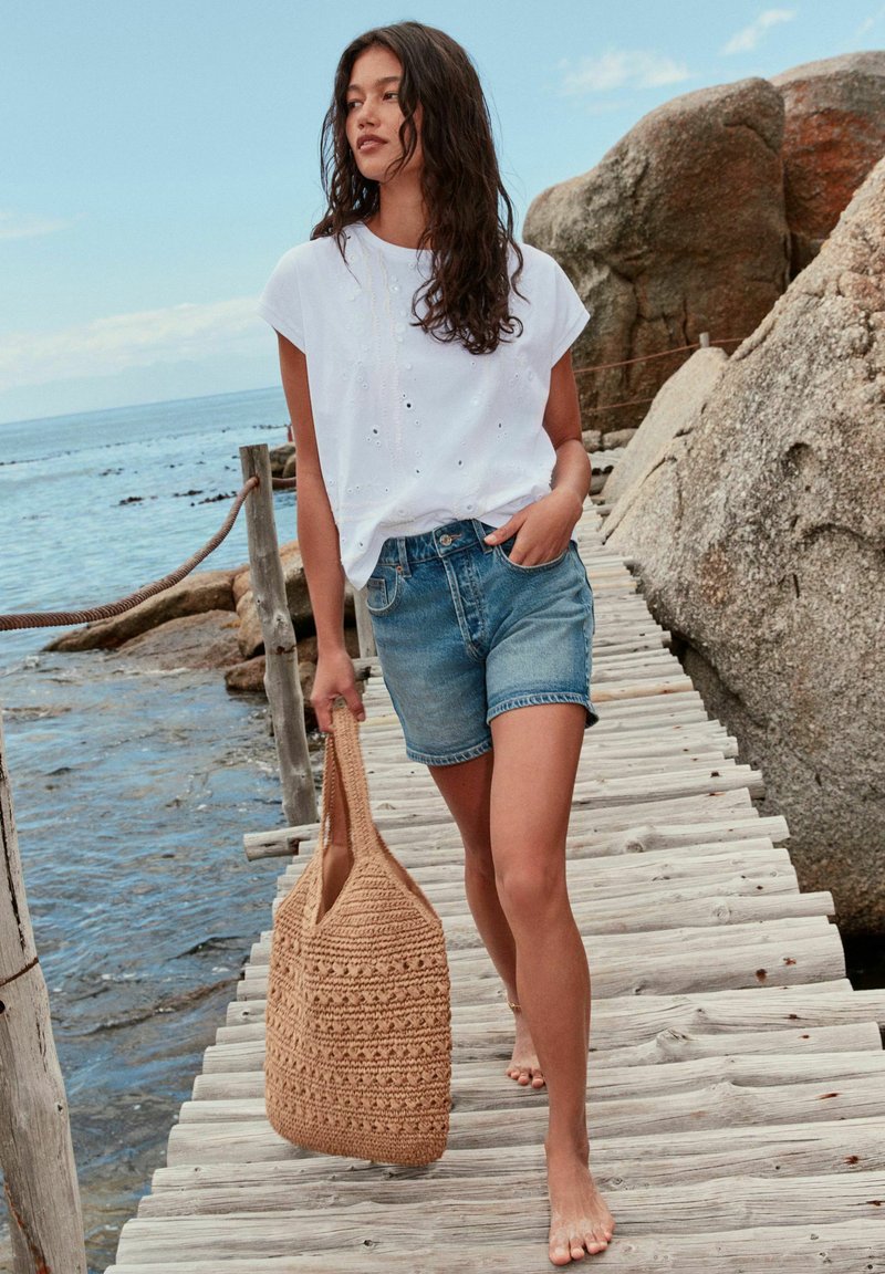 Femme en haut blanc à manches courtes et short en jean portant un sac tissé, marchant pieds nus sur une jetée en bois au bord d'une côte rocheuse et d'une mer calme.