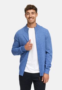 Cardigan en tricot bleu avec un col rond et une fermeture éclair intégrale. Il présente des poignets et un ourlet côtelés, porté par-dessus un t-shirt blanc.