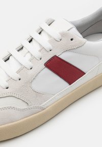 Sneaker bianco con accento in pelle rossa, tomaia in suede e sintetico, suola beige testurizzata e lacci piatti.