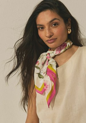 Frau mit langen dunklen Haaren, die ein cremefarbenes Oberteil, einen floralen Schal mit rosa und gelben Akzenten um den Hals gebunden und goldene Creolen trägt.